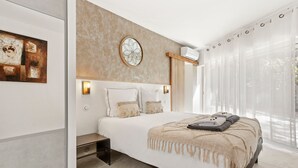 Een moderne slaapkamer met een groot bed, een hoofdeinde met een klok, een nachtkastje en uitzicht op de omgeving door dunne gordijnen.