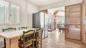 Een eetgedeelte met een houten tafel en stoelen, een koelkast en uitzicht door een raam.