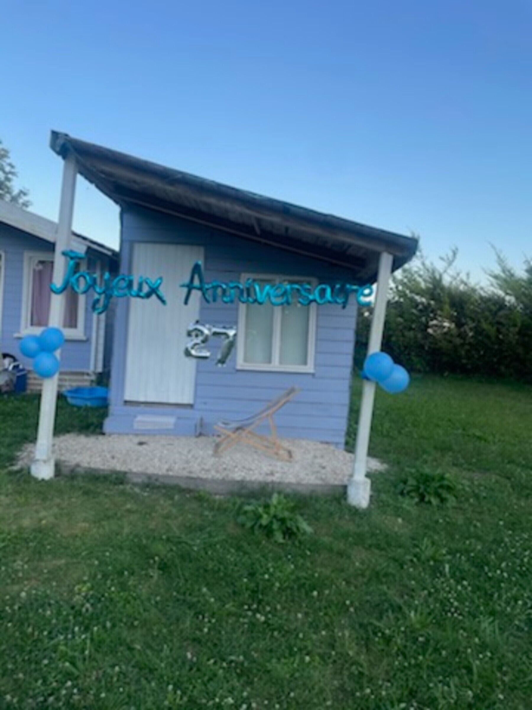 A small, single-story building with a blue exterior, a white door, and the words 'Joyeux Anniversaire' on the front.