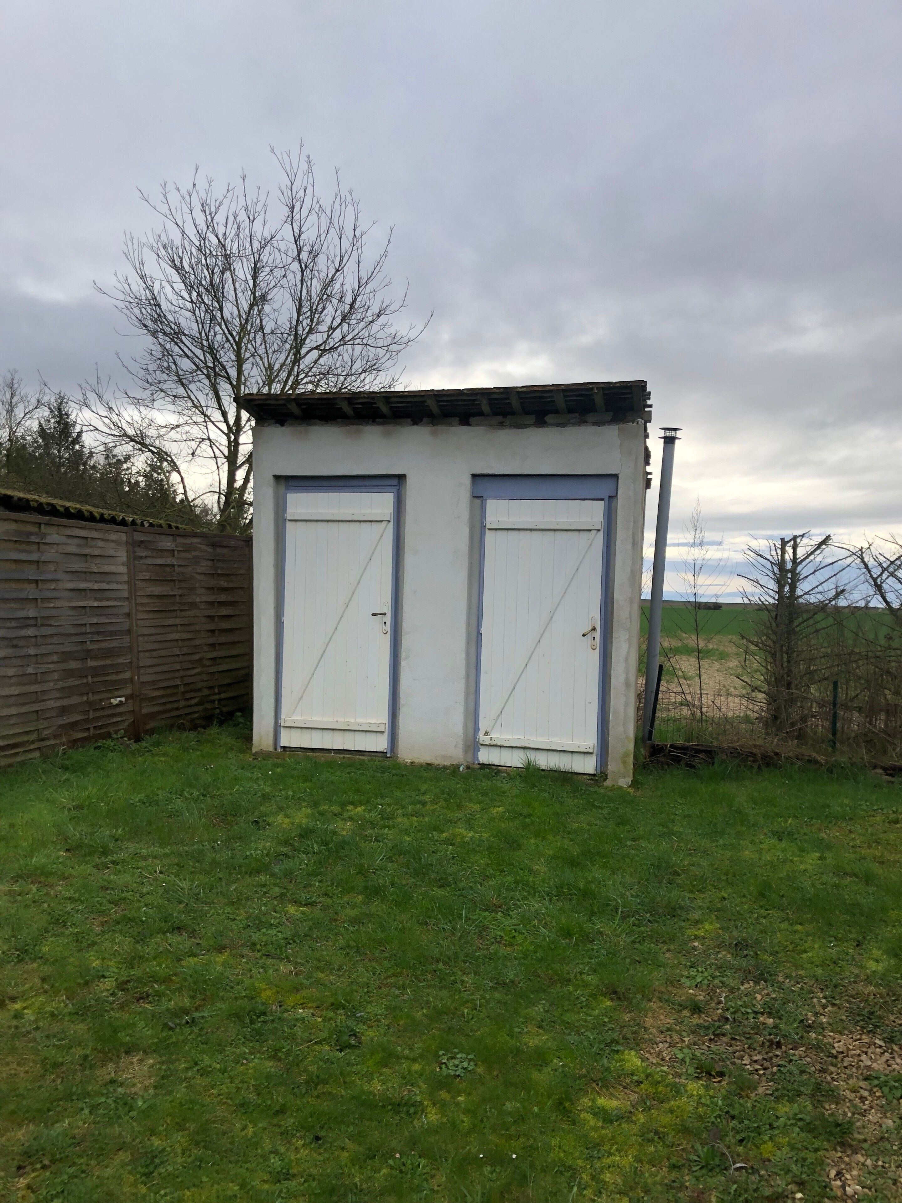 Een klein, laag gebouw met twee deuren, een schuin dak en een houten hek op de achtergrond.