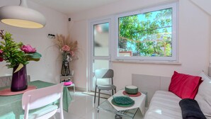 Un salon moderne et bien éclairé, comprenant un coin repas, un canapé agrémenté d’un coussin rouge, une table basse en verre et une fenêtre donnant sur la verdure.