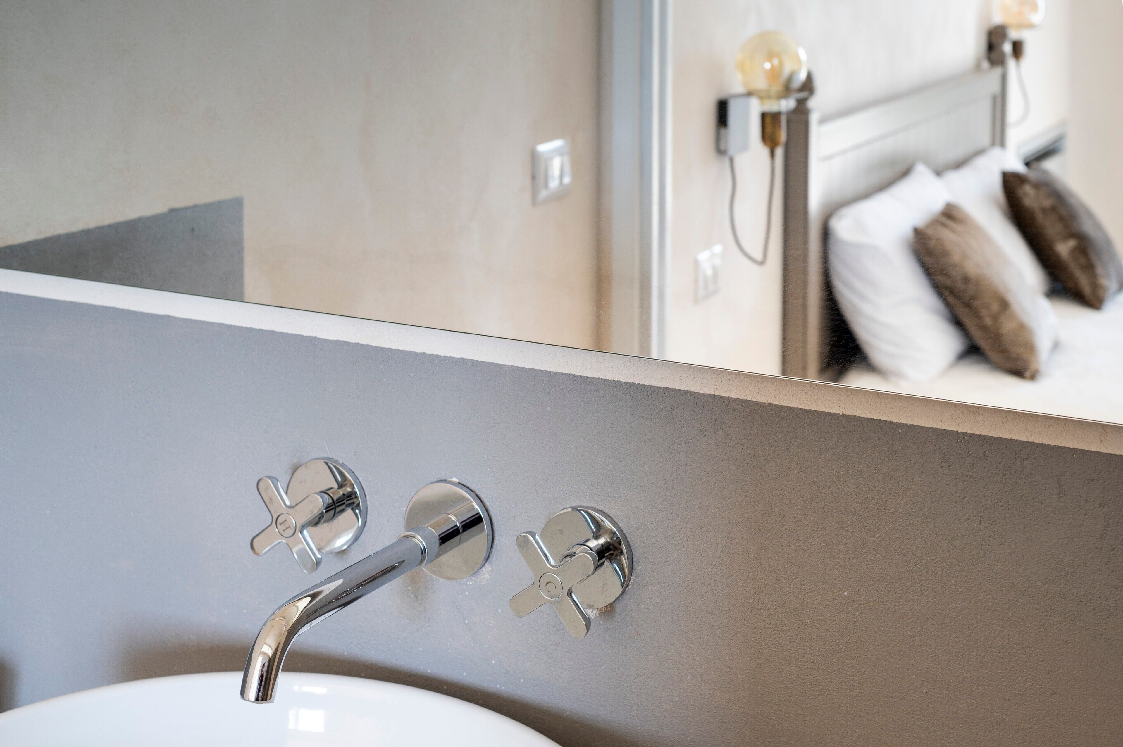 Un baño con un lavamanos moderno, grifería cromada y un espejo que refleja una cama con almohadas.