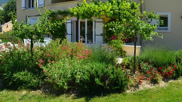 En tvåvåningsbyggnad med en balkong, omgiven av frodig grönska och blommande växter.