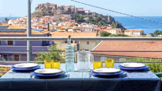 A table set with plates, glasses, and a pitcher, overlooking a coastal town with a prominent hill. - 'Ad Occhi Aperti - Castelsardo' Apartment with Sea View, Wi-Fi, and Air Conditioning (Castelsardo)