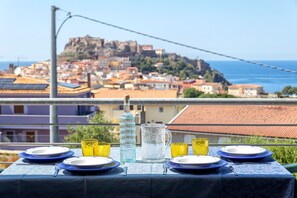 Mesa dispuesta con platos, vasos y una jarrón, con vista a un pueblo costero y una colina destacada.