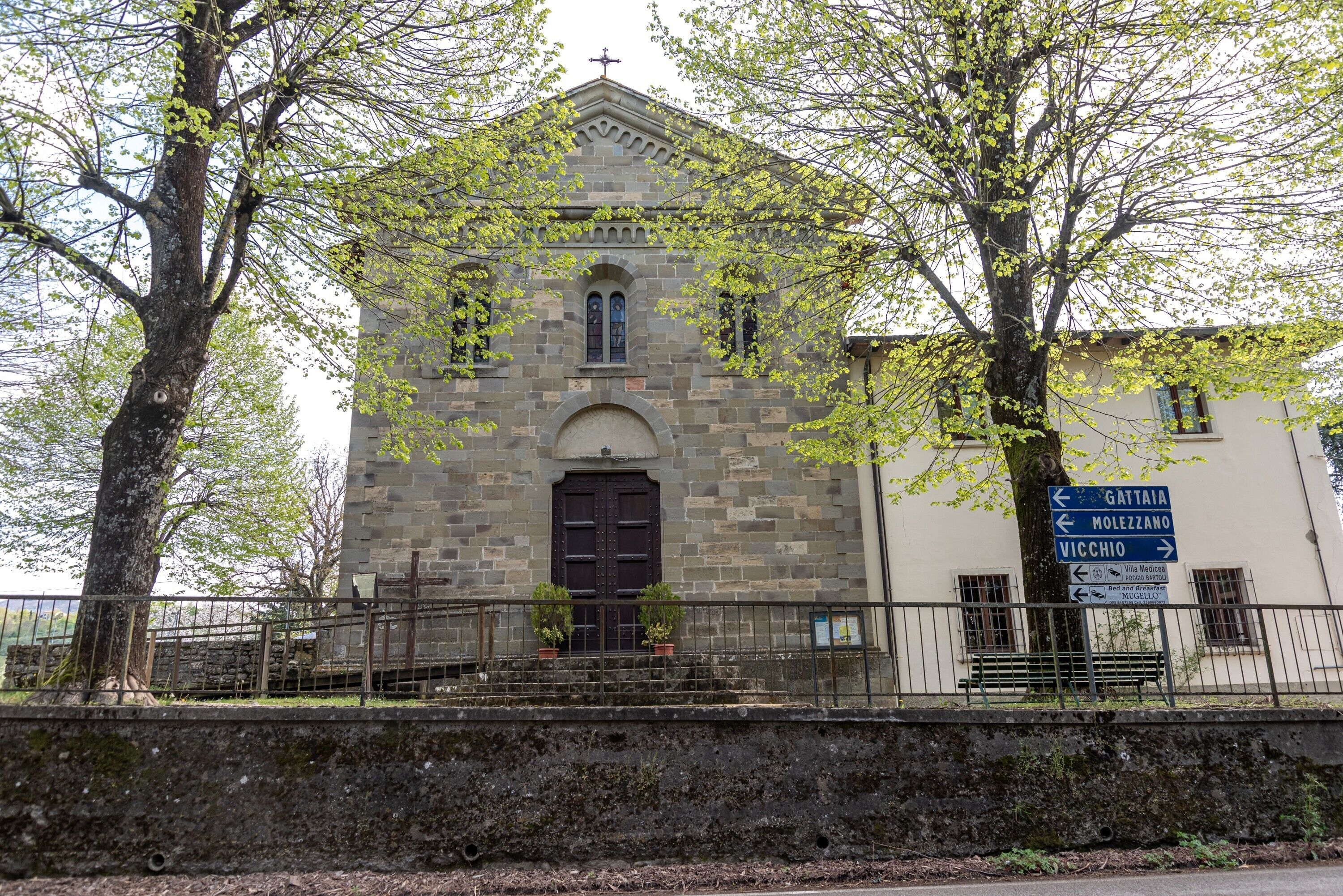 A stone church with a dark door and arched windows, surrounded by trees and a fence.