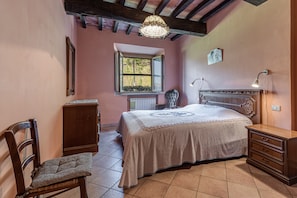 Un dormitorio con cama, una silla, cómoda, mesita de noche, lámpara, ventana y candelabro.