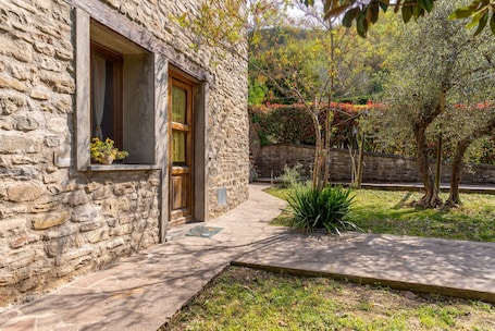A stone building with a wooden door and window, a paved pathway, and a small garden with plants and trees.