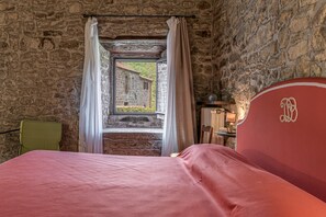 Una habitación con paredes de piedra, una cama con cabecera roja, una ventana con cortinas y vista a un edificio.