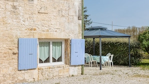 A stone building with blue shutters, a covered outdoor seating area, and a gravel ground. - Holiday Home 'La Rétaise' with Private Terrace and Garden (Romegoux)