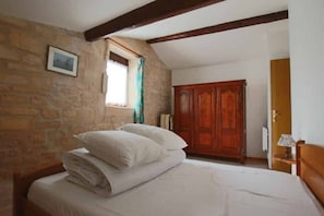 A bedroom with a bed, a wooden wardrobe, a window with curtains, and a stone wall.