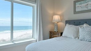 A bedroom with a large window overlooking a beach, a bed with white bedding, a nightstand with a lamp, and a framed picture on the wall.