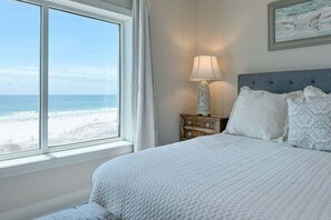 Un dormitorio con un ventanal que da a una playa, una cama con ropa de cama blanca, una mesita de noche con una lámpara y un cuadro en la pared.