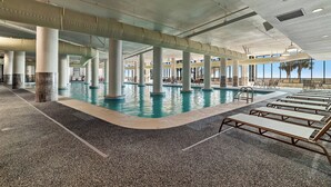 An indoor swimming pool with clear blue water, surrounded by lounge chairs and supported by large columns.