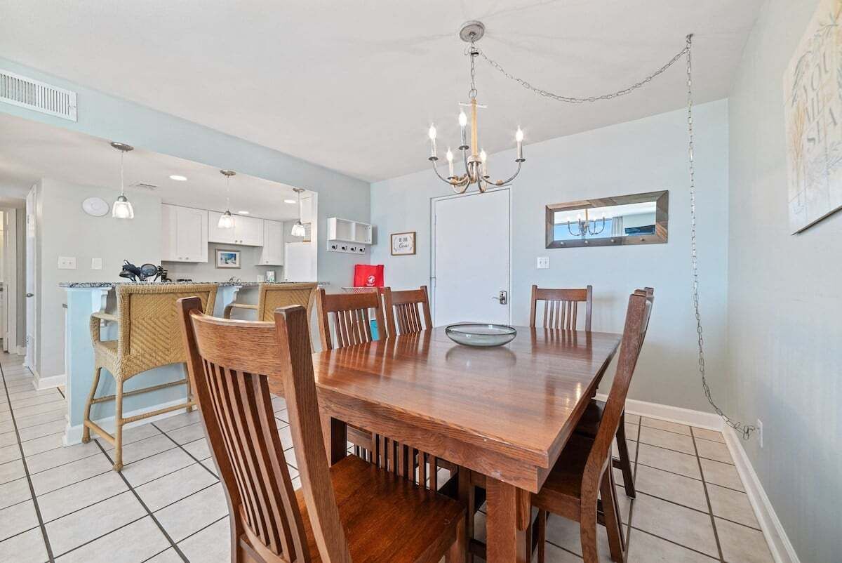 A dining area with a wooden table and chairs, a chandelier, and a kitchen in the background.