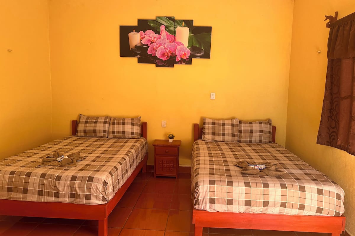 Una habitación con dos camas, cada una con colcha y almohadas a cuadros, una mesita de noche con lámpara, y un cuadro de flores y una vela colgados en la pared.