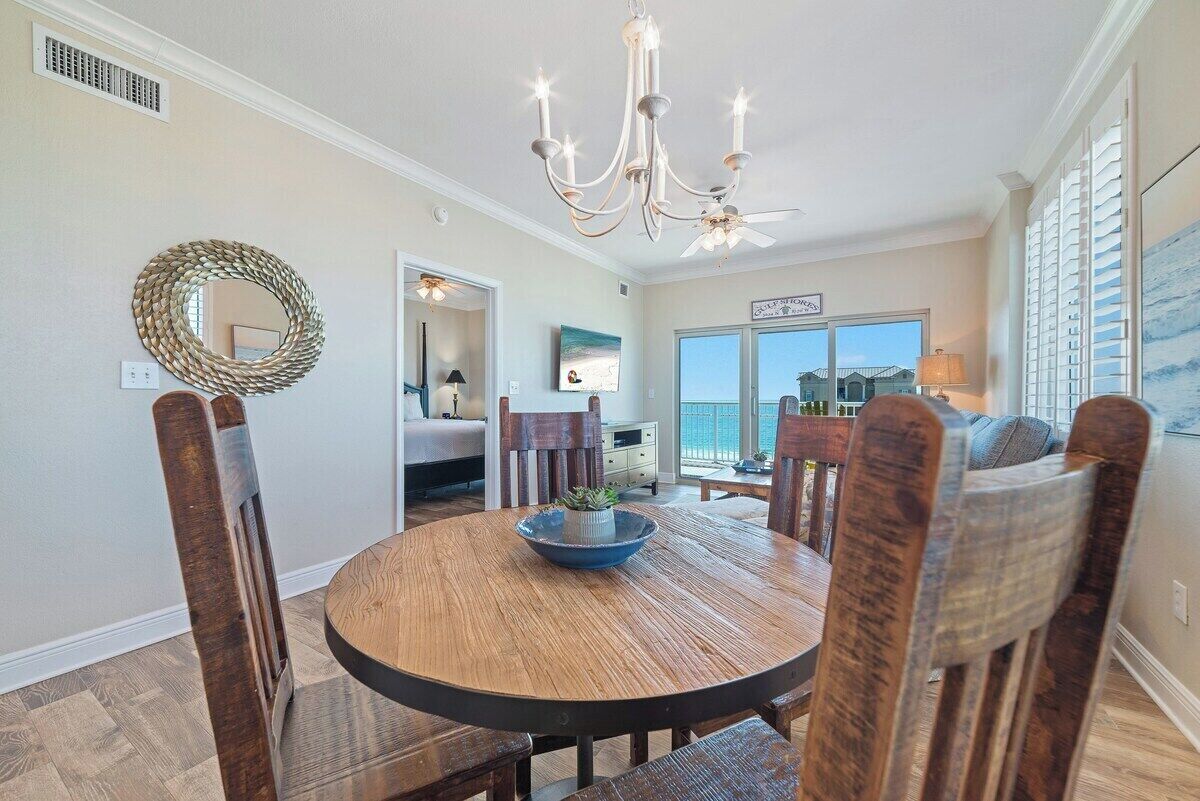 A dining area with a round wooden table, chairs, a chandelier, and a view of the ocean through large windows.