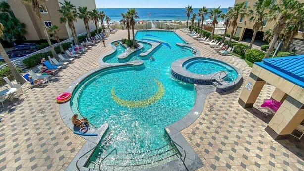 A swimming pool with a wavy design, surrounded by lounge chairs and palm trees, with a view of the ocean. - AMAZING CORNER UNIT, HUGE POOLS, BEACH VIEWS - Crystal Tower 809 (Gulf Shores)