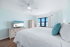 Een slaapkamer met een bed, een plafondventilator, een televisie, een raam met gordijnen en een dressoir.