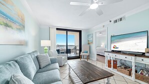 A living room with a gray sofa, a wooden coffee table, a TV on a white stand, and a view of the beach through a sliding glass door. - BEACHFRONT, FAMILY FRIENDLY, LOW FLOOR, OUTDOOR POOL (Orange Beach)