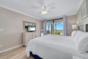 A bedroom with a large bed, a ceiling fan, a TV on a dresser, and a balcony with a view of palm trees and the sky.