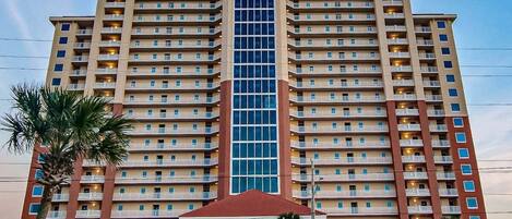 Un hôtel à plusieurs étages doté d’un ascenseur central à cage de verre, de nombreux balcons et d’une entrée surmontée d’un toit rouge.