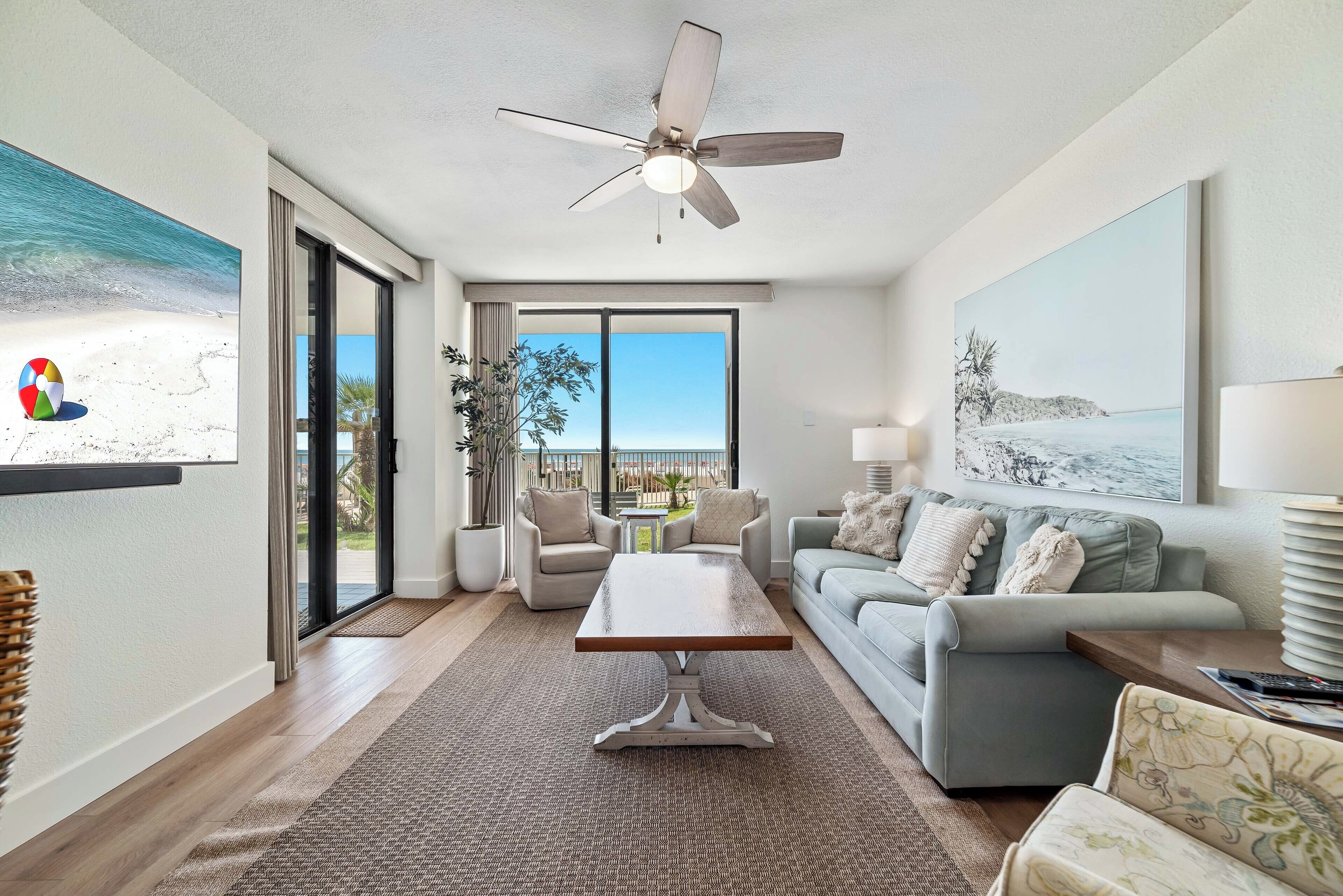 A spacious living room with a large window, a ceiling fan, and a view of the beach.