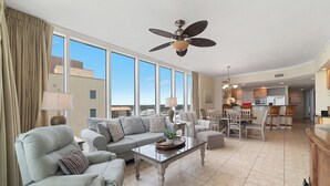 A spacious living area with a sofa, armchair, and coffee table, a dining area with chairs, and a kitchen in the background. - BEACHFRONT, LOW DENSITY CONDO, CORNER UNIT, CLOSE TO RESTAURANTS - The Colonnades 703 (Gulf Shores)