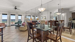 A spacious living area with a glass dining table surrounded by wooden chairs, a ceiling fan, and a view of the outdoors. - BEACHFRONT, FAMILY FRIENDLY, LOW FLOOR, INDOOR/OUTDOOR POOL (Orange Beach)