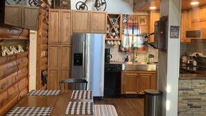 A log cabin interior with wooden walls, a dining table with checkered placemats, a refrigerator, a dishwasher, and a mounted deer head.