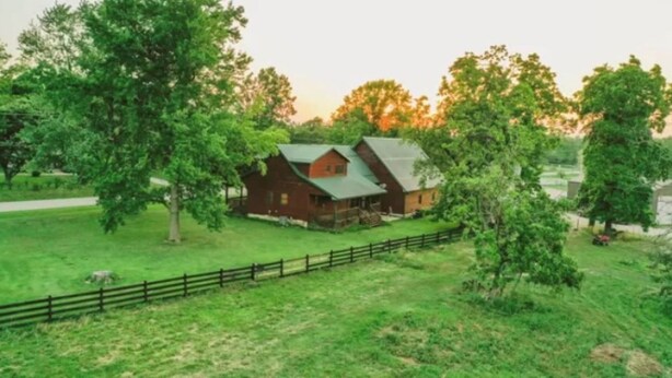 A rustic wooden cabin with a green roof, surrounded by lush greenery and a wooden fence.
