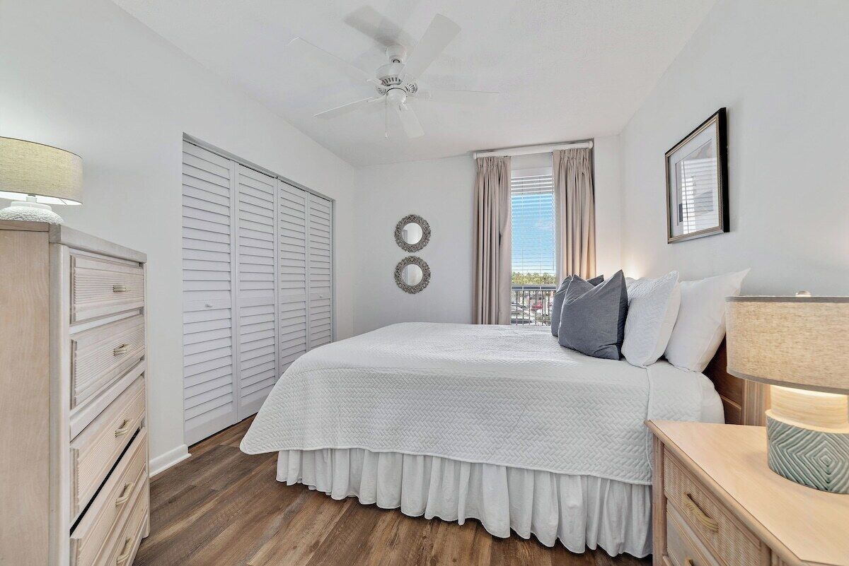 A bedroom with a bed, a ceiling fan, a window with curtains, a dresser, and a nightstand with a lamp.