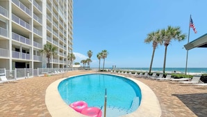 A swimming pool with a pool ladder, surrounded by lounge chairs and palm trees, with a building in the background.