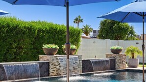 A pool area with a waterfall feature, surrounded by greenery and outdoor seating. - The Bridle is Right | Saltwater Pool | Backyard Games (Mesa)