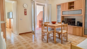 A compact kitchen with wooden cabinets, a dining table with chairs, a large window, and a view of a red brick building outside. - Casa Vacanza La Rosa (Giardini Naxos)