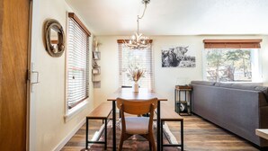 Un coin repas avec une table en bois, des chaises et un lustre. On aperçoit un canapé et une fenêtre avec des stores à l'arrière-plan.