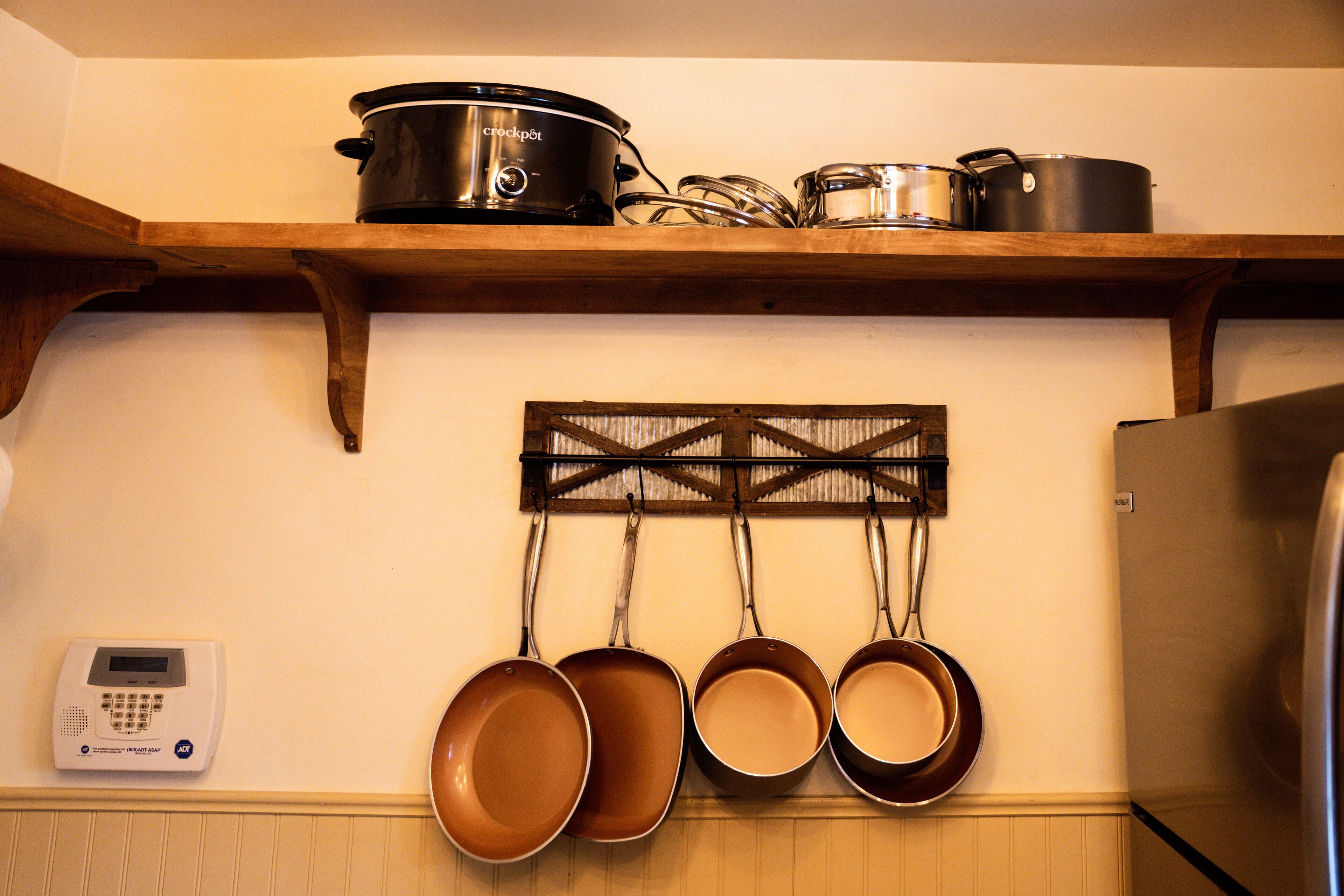 Une cuisine avec une étagère en bois où sont rangés les ustensiles de cuisine, un porte-casseroles fixé au mur et un appareil numérique accroché au mur.