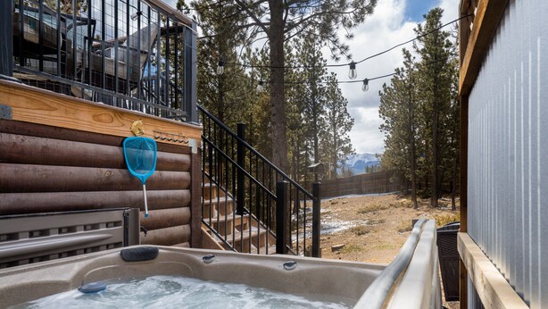 Un espace avec un bain à remous, doté d’une terrasse en bois, d’une rambarde en métal et offrant une vue sur une zone boisée, avec des montagnes en arrière-plan.