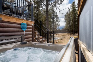 A hot tub area with a wooden deck, metal railing, and a view of a forested area with mountains in the background. - The Hideout (Woodland Park)