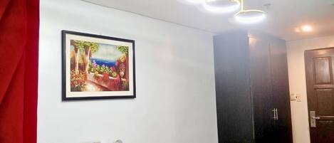 A hotel room with a bed, a desk with a chair, a framed painting, a black wardrobe, and a ceiling light fixture.