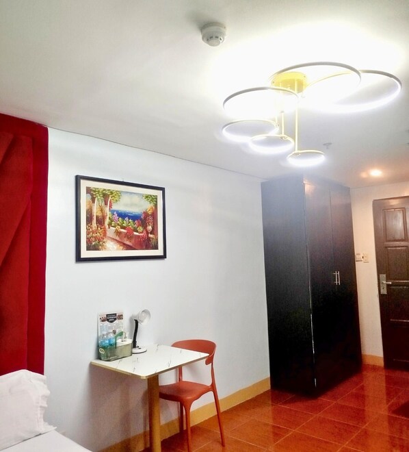 A hotel room with a bed, a desk with a chair, a framed painting, a black wardrobe, and a ceiling light fixture. - Haeinsa Condotel (Quezon City)