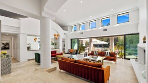 A spacious living area with a large sofa set, a coffee table, and a dining table. There are multiple windows providing natural light and views of the outdoors.