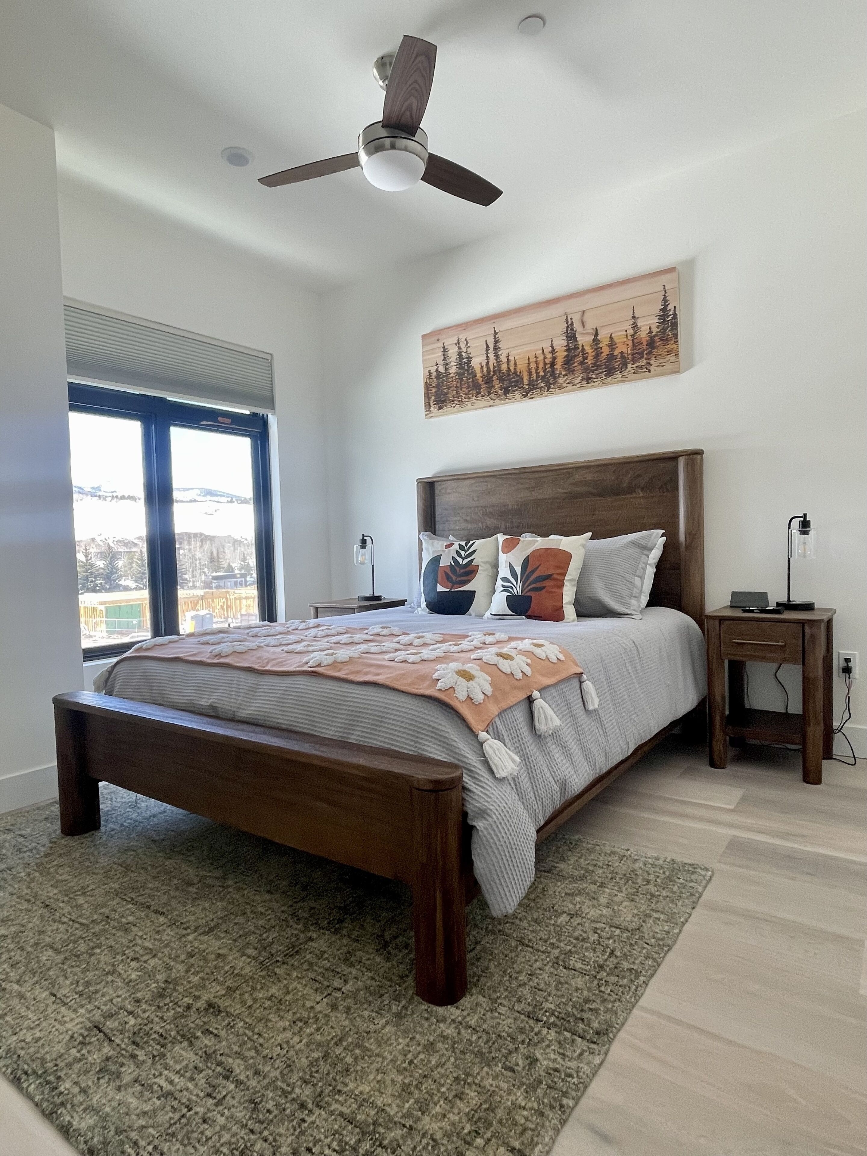 A bedroom with a bed, a ceiling fan, a nightstand, a bedside table, and a window with a view.