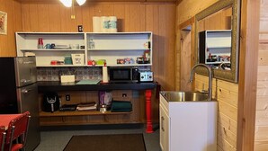 A compact kitchen with wooden walls, a ceiling fan, a refrigerator, a microwave, a sink, and a small dining area with a red chair.