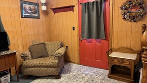 A cozy room with a patterned armchair, a red door, a small wooden table, and a wall-mounted light fixture.