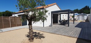 A modern outdoor patio area with a wooden deck, a covered seating area, and a small tree. - "La balise jaune", new house on the edge of the forest, ideal for families! (Saint-Jean-de-Monts)