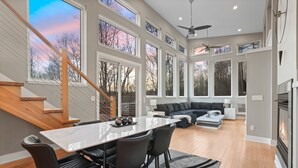 A spacious living area with a large window, a staircase, a dining area with a table and chairs, and a fireplace. - The Fairchild Haus | 30 Acres, Sauna, EV Charger (Logan)