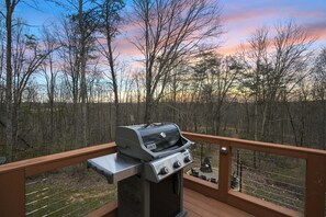 Una parrilla Weber Spirit sobre una terraza de madera con vista a un área boscosa y un atardecer al fondo.