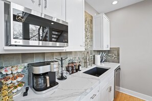 A modern kitchen with white cabinetry, a built-in microwave, a coffee maker, and a sink with a stone backsplash.
