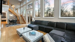 Ein großzügiger Wohnbereich mit einer großen, schwarzen Eckcouch, einem Couchtisch und einem Essbereich mit Tisch und Stühlen. Eine Holztreppe führt zu einer oberen Ebene.